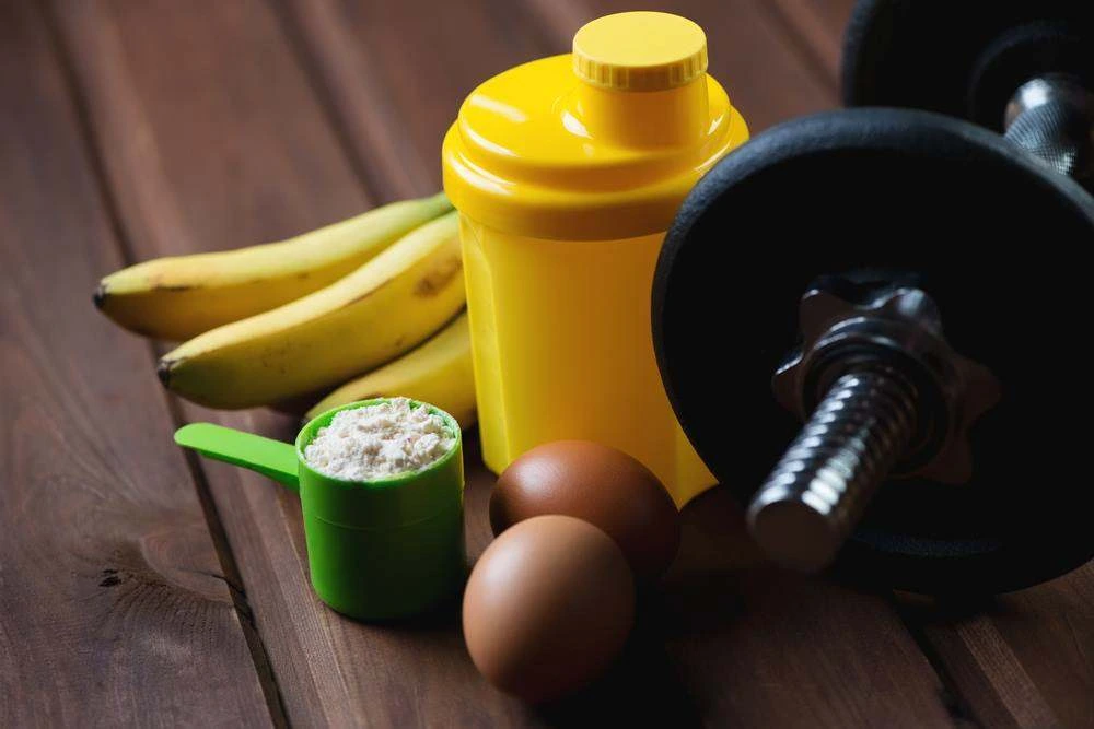 Protein powder, a shaker, bananas and eggs beside a dumbbell